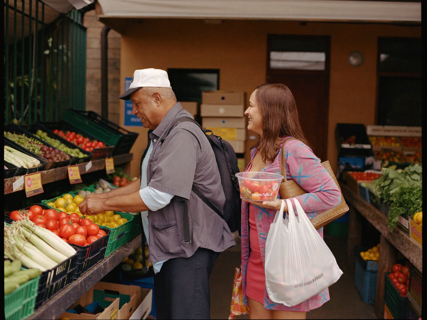 An older man choosing tomatoes at a market stall while his companion holds the bag